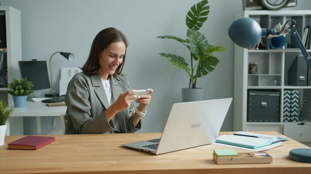 A woman in a home office using a smartphone while working on a laptop.
